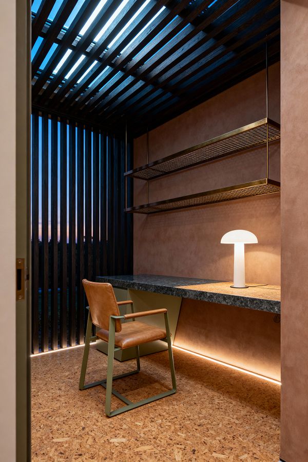 A compact contemporary home office with a built-in dark stone desk set against warm terracotta walls. Two brass-framed mesh shelves hang from slender rods above a glowing white dome table lamp. A tan leather chair with a sage-green metal frame sits on cork flooring. The ceiling and adjacent window are wrapped in dark vertical timber slats that filter narrow bands of evening light, while an LED strip illuminates the wall and floor beneath the desk.