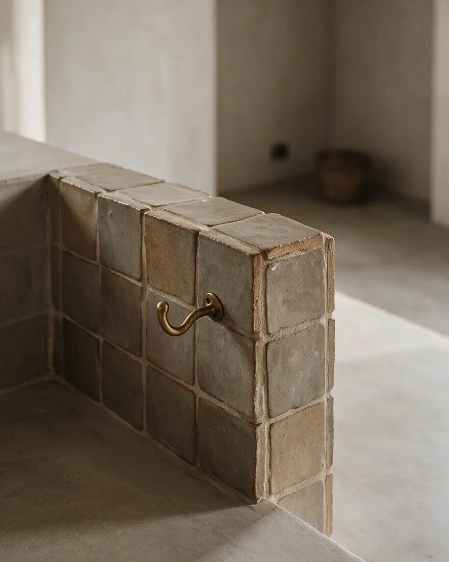 A close-up view of a bath or shower wall finished in small square handmade tiles with uneven edges and earthy tones. A simple curved brass hook is mounted on the tiled surface, while the background shows a quiet minimalist interior in soft beige and gray light.