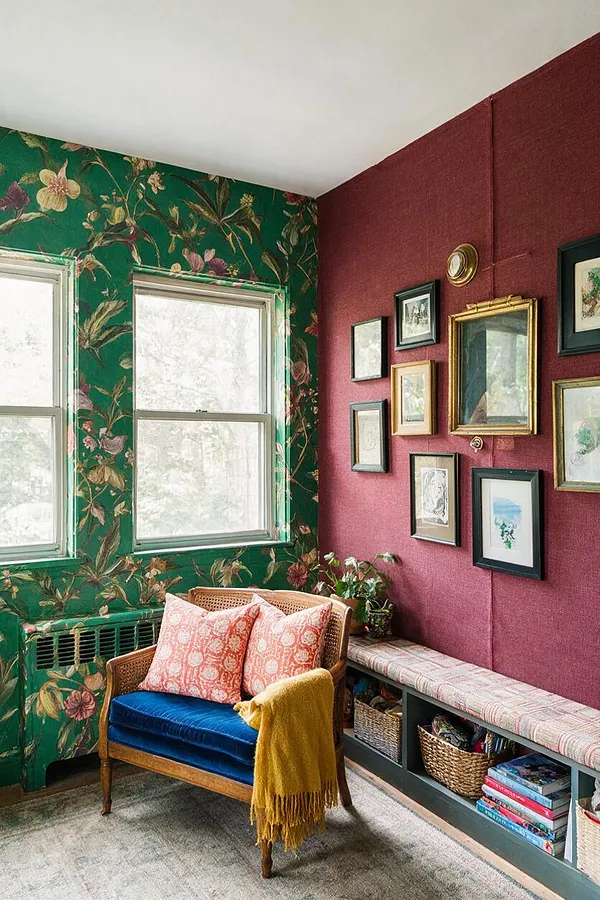 A colorful sunroom with green floral wallpaper, burgundy textured walls, and a cluster of framed artwork. A woven rattan chair with a deep blue velvet seat, coral patterned pillows, and a mustard throw sits beside a built-in bench with storage baskets and books.