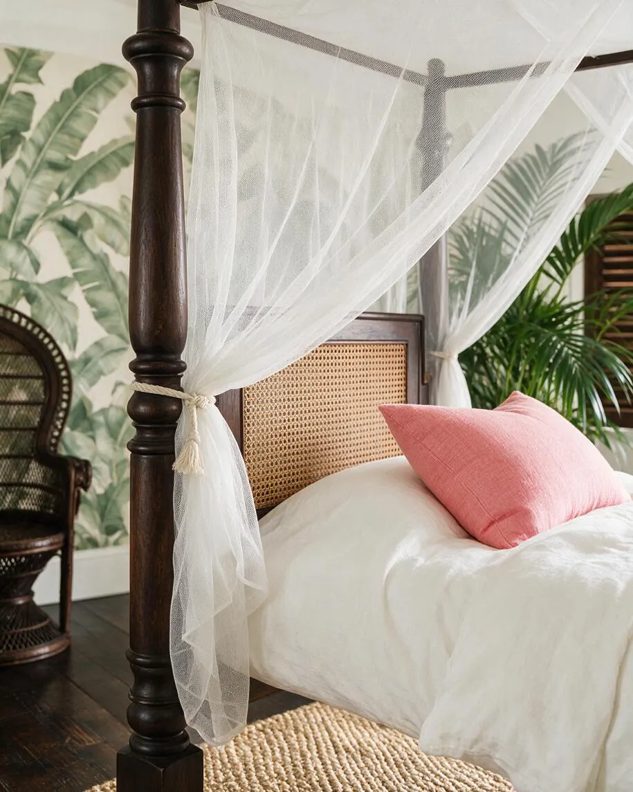 A tropical-style bedroom featuring a dark wood four-poster bed with sheer white canopy drapes, a woven cane headboard, white bedding, and a soft pink accent pillow. Palm-leaf wallpaper and a woven chair in the background reinforce the relaxed, resort-like feel.