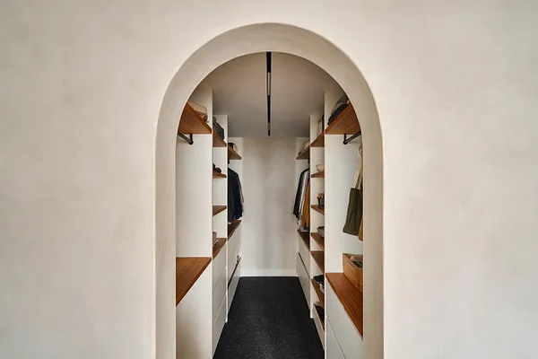 A narrow modern walk-in closet is framed by a soft arched doorway and lined with white built-in cabinetry. Warm wood shelves and hanging rods run along both sides, with a dark floor runner and a minimalist ceiling light creating a clean, streamlined look.
