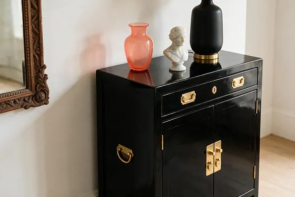A glossy black cabinet with brass hardware stands against a light wall, styled with a coral glass vase, a small white bust, and a matte black vase on top. An ornate carved mirror frame is partially visible on the left, adding to the eclectic mix of finishes and objects.