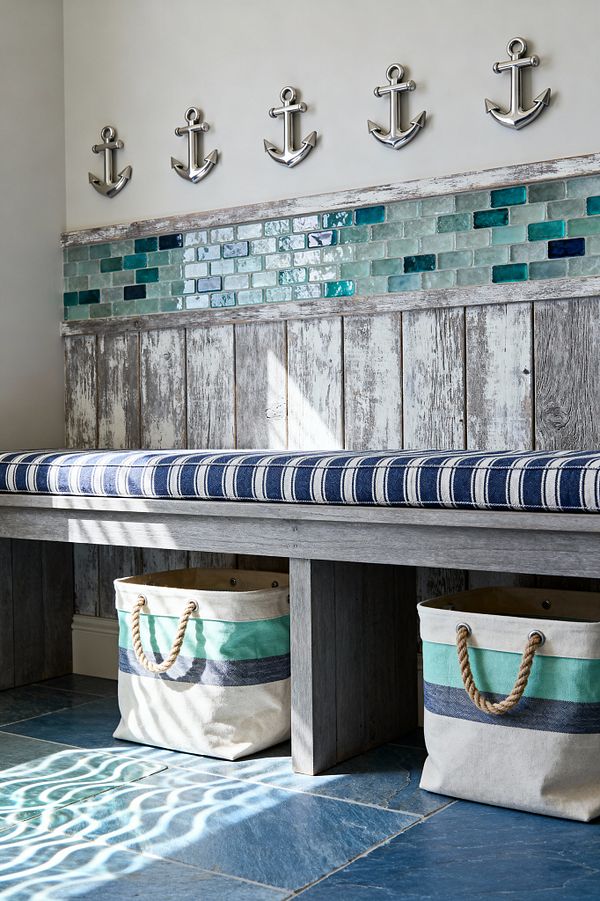 A coastal mudroom features a weathered gray wood bench with a navy and cream striped cushion. Behind it are reclaimed wood planks and a band of glossy glass subway tiles in aqua, teal, and deep blue, topped by five polished metal anchor hooks. Blue stone floor tiles reflect bright, rippled sunlight. Two canvas totes with seafoam and navy bands and rope handles sit beneath the bench for storage, reinforcing the nautical look.