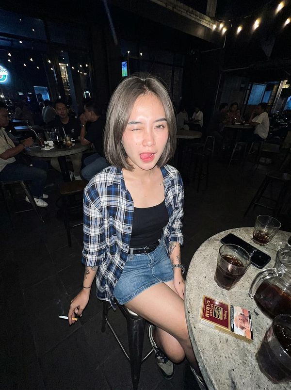 A young woman is sitting at a bar, playfully winking at the camera while holding a cigarette.