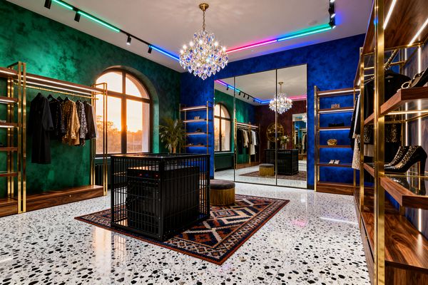 Large room styled as a maximalist home gym and dressing space with emerald and cobalt textured walls, floor-to-ceiling mirrors, and arched windows at sunset. Brass and walnut shelving lines the perimeter; a crystal chandelier and RGB ceiling strips illuminate white terrazzo flooring with black chips, a patterned rug, a velvet fringed ottoman, and a black metal cage in the center with garments and shoes displayed.