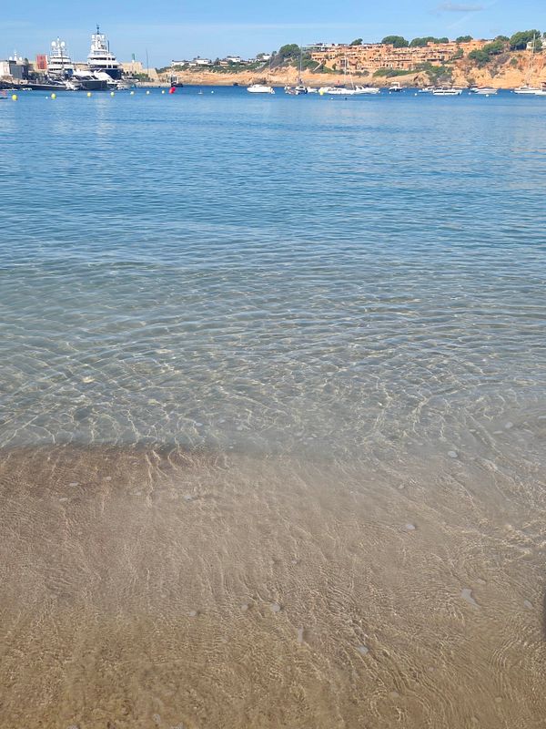 Clear shallow beach water with visible sandy bottom, yachts, sailboats, and coastal buildings in the background under a blue sky.