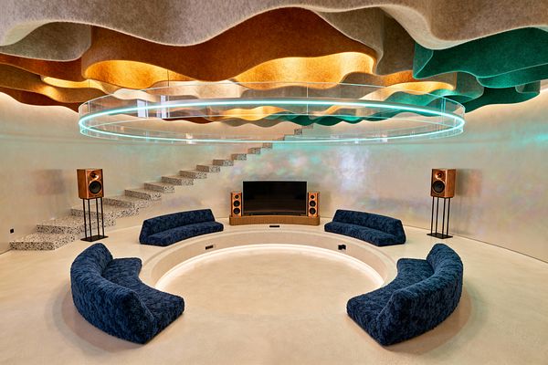 A futuristic media room with a circular, sunken seating pit framed by LED strip lighting and four curved navy boucle sofas. A large flat-screen sits on a curved wood console, flanked by multiple wood-cased speakers, while a terrazzo floating staircase rises along the left wall. Above, an undulating acoustic felt ceiling in tan, orange, gray, and teal is highlighted by a suspended double-ring acrylic light, casting a cool glow across pearlescent plaster walls and a pale microcement floor.