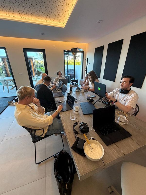 A group of six individuals working on laptops around a modern wooden table in a well-lit room with large windows and black acoustic panels on the wall.