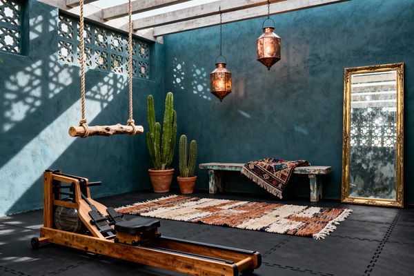 Bohemian-inspired home gym with deep teal plaster walls beneath a slatted pergola casting patterned shadows. A solid-wood water rowing machine rests on black rubber tiles beside a rust-and-ivory shag rug; hammered copper lanterns hang over a distressed turquoise bench topped with a kilim throw, while terracotta potted cacti, a rope swing with a driftwood seat, and a large antique gold-framed mirror lean against the wall.