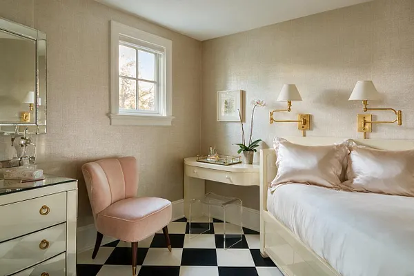 A stylish guest bedroom with a cream bed dressed in white bedding and blush satin pillows, paired with gold swing-arm wall lamps. The room features a black-and-white checkered floor, a pink velvet accent chair, a mirrored dresser, and a small cream desk under a window, creating a polished Hollywood Regency look.