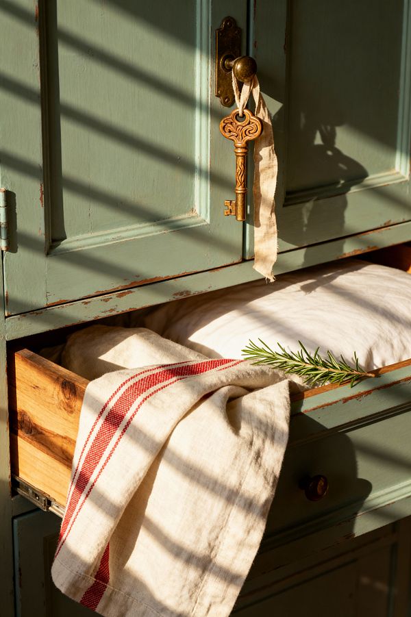 Close-up of a distressed sage-green armoire with paneled doors and a brass escutcheon, its round knob holding an ornate brass key tied with a linen ribbon. An open lower drawer in knotty pine reveals a white linen cushion; a natural flax grain-sack cloth with red stripes drapes over the edge beside a sprig of rosemary. Slanted sunlight casts narrow shadows, emphasizing the chippy paint, warm wood, and aged metal details.