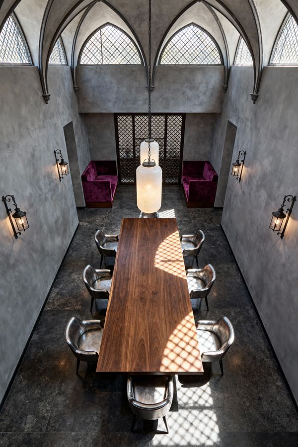 A Gothic-style dining room with a soaring ceiling of ribbed, pointed vaults and high leaded-glass windows. A long walnut table sits at center, surrounded by silver-toned leather chairs, with wrought-iron lantern sconces on gray limewashed plaster walls and a dark stone floor. Two chain-hung cylindrical alabaster pendants hang above, and plum velvet banquettes sit in rear niches beside a decorative iron screen, with patterned light falling across the space.