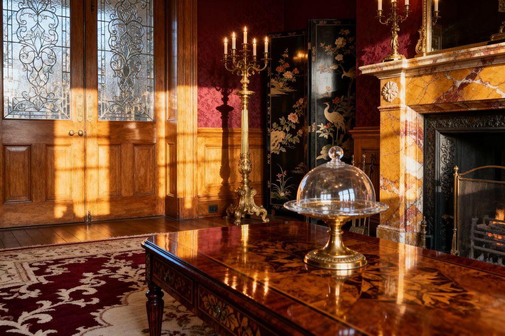 A Victorian living room bathed in late-afternoon sun features carved oak paneling and double doors with ornate leaded glass. A gilded candelabrum glows beside a black lacquer Chinoiserie screen. The fireplace has a golden Siena marble mantel and a black cast-iron insert with a small fire. A burgundy-and-ivory patterned rug anchors an inlaid marquetry coffee table topped with a glass cloche on a brass stand.