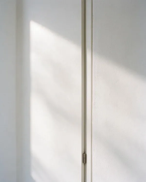 A close-up of a minimalist interior detail showing smooth white wall panels divided by a narrow vertical trim line. Soft sunlight creates a triangular glow and faint shadow across the surface, emphasizing the clean lines and subtle texture.