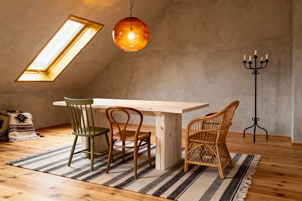 An attic dining nook with sloped greige plaster walls and a skylight centers on a pale pine block‑leg table. Three mismatched chairs—a sage painted spindle chair, a bentwood chair with a natural rush seat, and a woven rattan armchair—sit on a cream and charcoal striped flatweave rug. An amber glass globe pendant casts warm light above, while a black wrought‑iron candelabra stands to the right on honey‑toned pine floors.