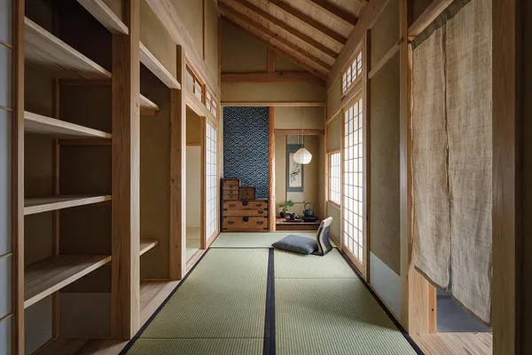 A narrow Japanese Zen-style closet or dressing room with open wooden shelving on the left, tatami mat flooring, and shoji screens lining the walls. Natural linen curtains hang on the right, and warm wood beams and trim create a calm, minimalist atmosphere.