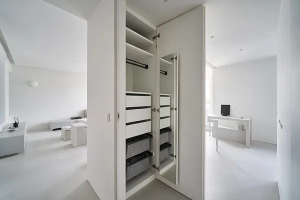 A minimalist built-in closet with a tall hinged mirrored door, open white shelving, a black hanging rod, and stacked white drawers with dark trim. The closet is set within a bright white room with clean lines and sparse furnishings, creating a calm, airy atmosphere.