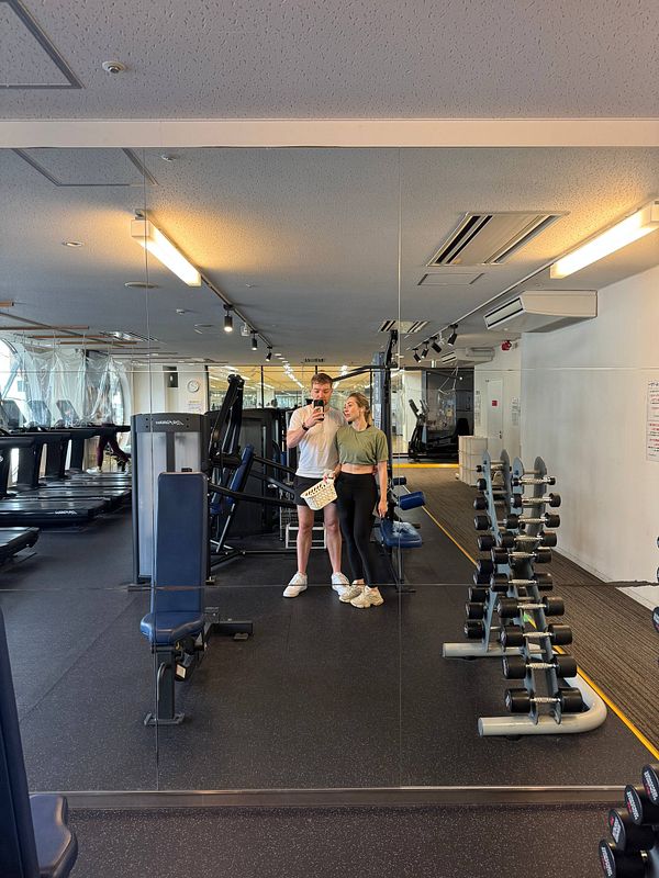 A couple poses for a selfie in a gym with various workout equipment in the background.