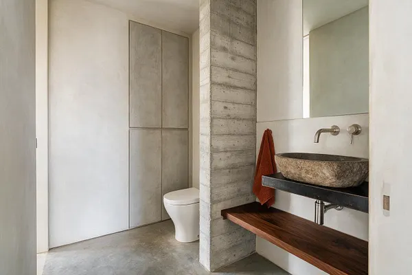 A compact powder room with a Brutalist aesthetic, featuring a textured concrete partition, a stone vessel sink on a dark floating shelf, a warm wood counter below, and a white toilet tucked into a recessed alcove. Large mirror, matte wall surfaces, and minimal fixtures create a clean, sculptural look.