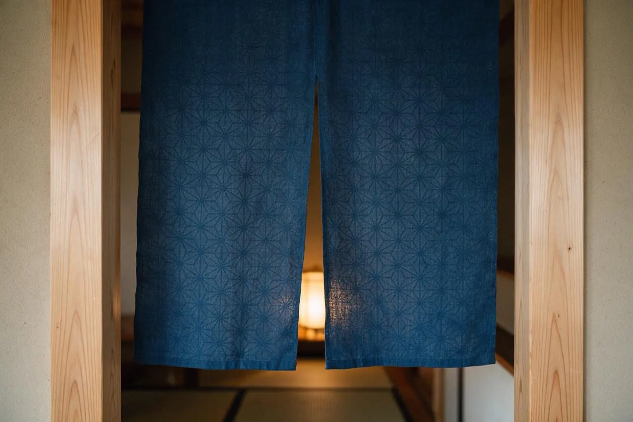 A Japanese Zen-style doorway framed in pale natural wood with textured blue noren curtains hanging in the center. A warm lantern glow is visible behind the fabric, with a calm tatami-lined interior beyond.