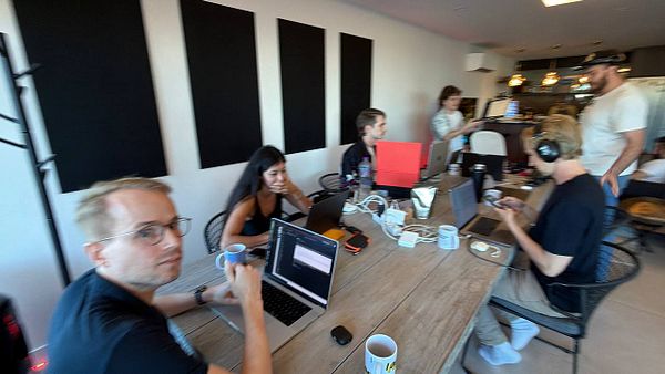 A group of seven people working and interacting in a modern coworking space around a large wooden table with laptops and coffee mugs.