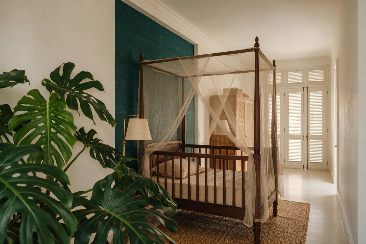 A tropical-style nursery with a dark wood canopy crib draped in sheer mosquito netting, set beside a teal accent wall and a woven rug. Large monstera plants fill the foreground, while a wicker storage cabinet and bright shuttered doors add warmth and natural texture.