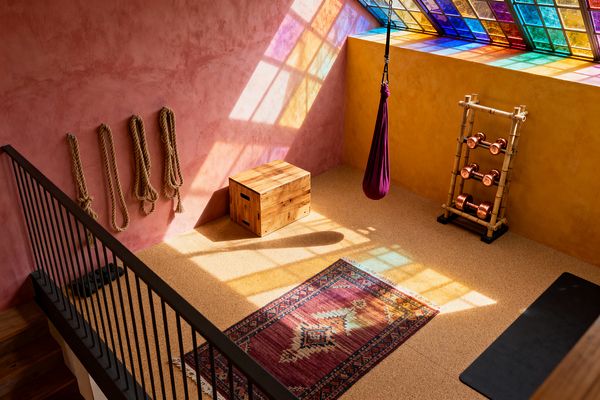 Bohemian-style home gym with dusty pink and mustard plaster walls, a cork floor, and a patterned kilim rug. Colored light from a stained-glass skylight falls across a plum aerial yoga hammock, a natural wood plyo box, and jute ropes hung on the wall. A bamboo rack holds copper dumbbells beside a black yoga mat, with a simple black stair railing in the foreground. The room feels bright, warm, and handmade.