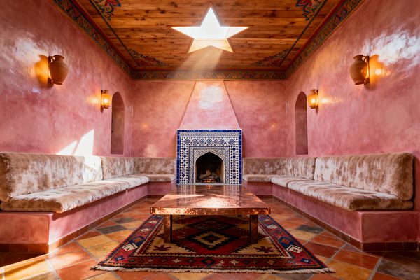 A Moroccan-style living room with glossy pink tadelakt walls features built-in banquette seating upholstered in cream velvet along both sides. A hammered copper coffee table sits on a red Berber kilim over terracotta tiles, facing a blue-and-white zellige-tiled fireplace with a triangular plaster hood. A wooden ceiling with a painted border is pierced by a star-shaped skylight that sends sunbeams across arched niches and warm clay sconces.