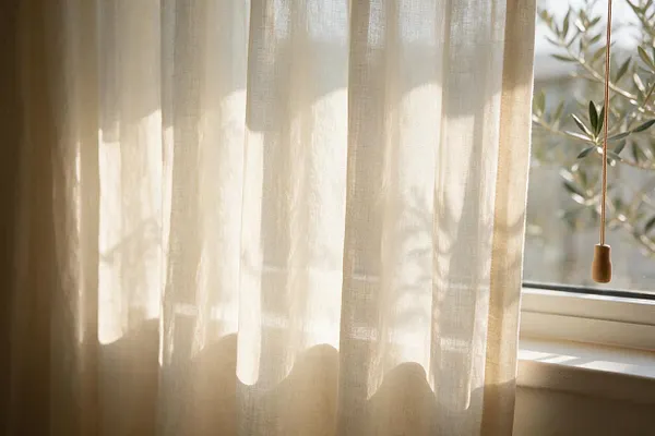 A close view of translucent beige linen curtains hanging beside a sunlit window. Soft shadows from nearby foliage fall across the fabric, and a pull cord hangs at the right, reinforcing the calm, nature-inspired feel.