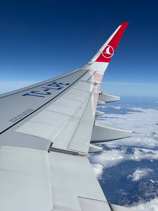 View from an airplane window showing a Turkish Airlines wing flying over mountains and clouds under a clear blue sky.