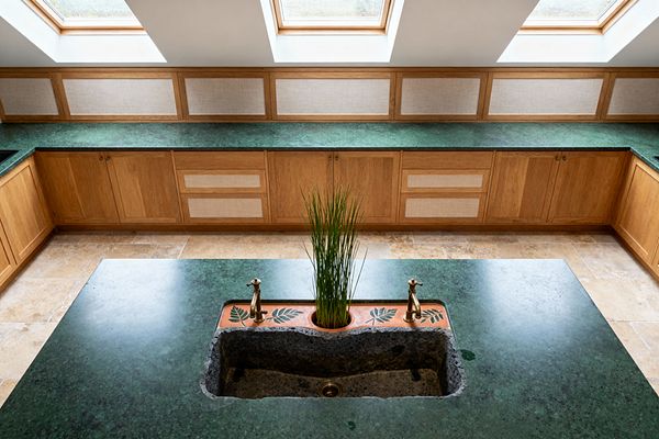 A bright kitchen with U-shaped oak cabinetry and deep green stone countertops sits beneath three large skylights. The island features a rough stone trough sink flanked by brass taps, set into a terracotta deck stamped with leaf motifs and a circular planter holding tall grass-like greenery. Light beige textured wall panels run above the counters, and beige limestone floor tiles complete the warm, natural look.
