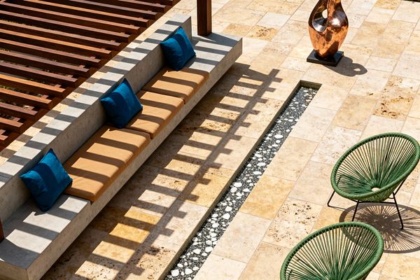 Sunlit patio with a long cast-concrete built-in bench under a slatted wood pergola that throws striped shadows across the seating. The bench has camel-colored seat cushions with deep teal throw pillows. The ground is paved in warm beige travertine tiles with a narrow linear channel filled with dark pebbles and white stones. Two round, green cord-woven lounge chairs sit on thin black metal legs, and a burnished copper sculpture stands on a small black plinth nearby.
