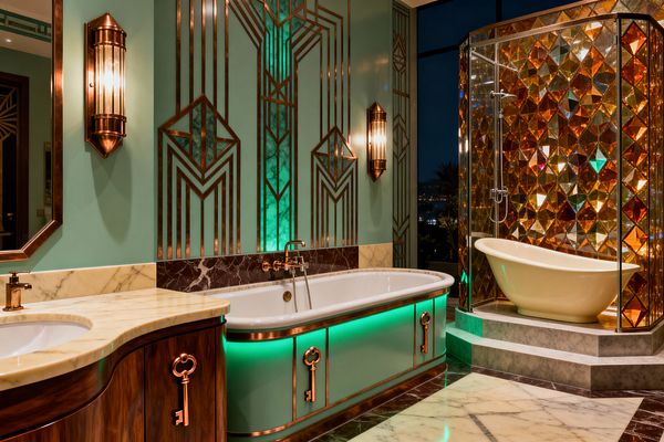 A luxurious Art Deco bathroom features mint-green paneled walls with copper geometric inlays, a white oval tub with copper trim and green underlighting, and a curved vanity in rich wood with a cream marble top and key-shaped pulls. Fluted copper sconces flank the tub, while a glass shower enclosure glows with amber, faceted glass; a cream slipper tub sits on gray stone steps above a marble floor, with a nighttime city view beyond.