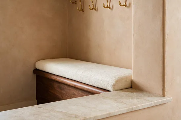 A compact mudroom nook with smooth beige plaster walls, a cream cushioned bench, a rich wood base, and brass wall hooks above. A light stone ledge in the foreground adds a refined natural finish to the calm, built-in seating area.