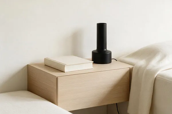 A minimalist bedroom vignette with a light oak floating nightstand against a white wall. A small cream-covered book and a black cylindrical lamp sit on top, with soft white bedding draped around the bedside.