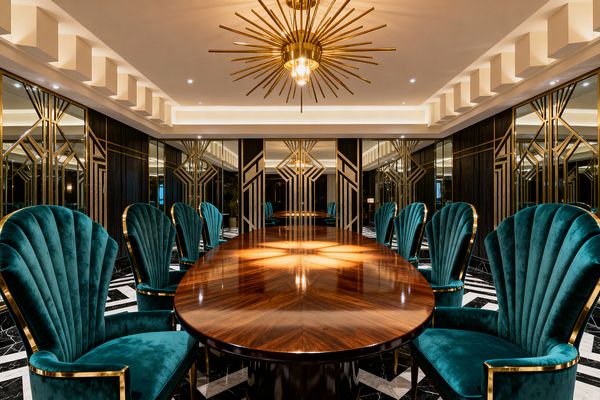 A luxurious Art Deco dining room with a long oval walnut-veneer table reflecting a brass sunburst chandelier. Deep teal shell-back velvet chairs with brass edging line both sides; walls combine dark wood paneling and mirrored panels with geometric gold inlays, and a black marble floor with white veining and a bold border anchors the room beneath a stepped ceiling with warm recessed cove lighting.