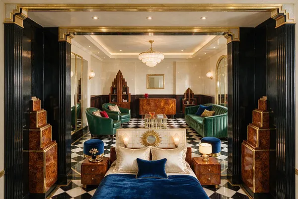 A luxurious Art Deco studio apartment interior with a black-and-white checkered floor, glossy black columns, and gold trim framing the space. The room features green velvet sofas, a blue velvet bed in the foreground, a crystal chandelier, mirrored and wood-paneled accents, and rich warm wood furniture that gives the room a dramatic, vintage-inspired elegance.