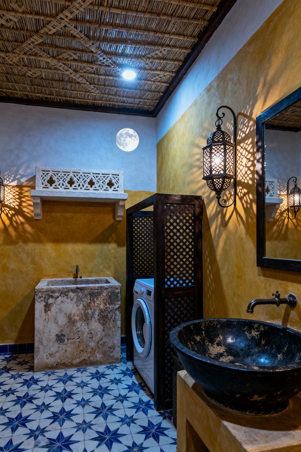 A Moroccan-inspired laundry room features ochre plaster walls and blue-and-white star-pattern floor tiles. A weathered stone wash trough sits beneath a carved plaster shelf, while a front-loading washer is tucked behind a dark wood lattice screen. On the right, a black stone vessel sink with a wall-mounted spout sits on a masonry counter. Perforated metal lanterns cast patterned light, and a woven reed ceiling and small round window with a moonlit view complete the space.