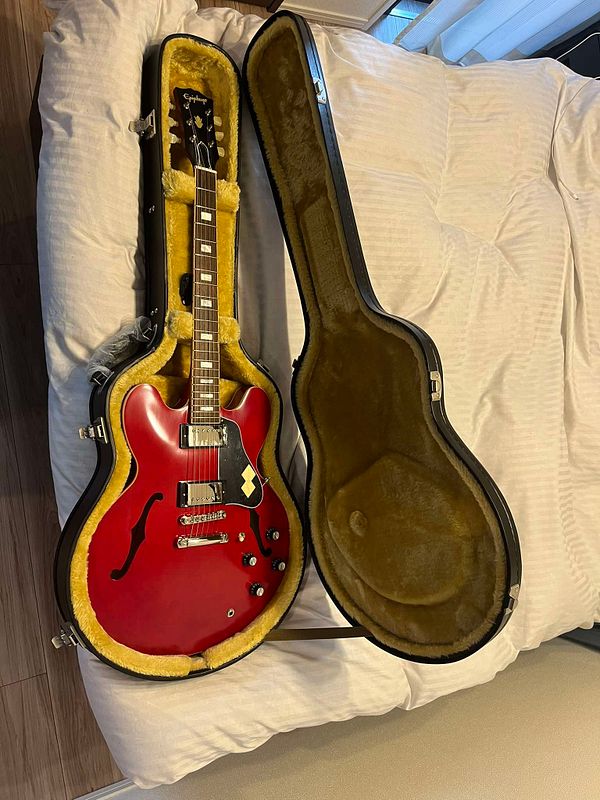 A red electric guitar is displayed in its open case on a bed.