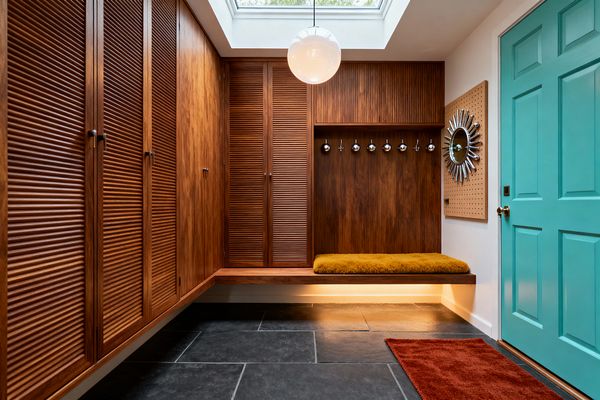 A mid-century mudroom features teak louvered cabinets along the left wall and corner, a floating wood bench with a mustard shag cushion and recessed lighting, and a wood back panel fitted with chrome ball coat hooks. The floor is large charcoal slate tile with a burnt orange runner. A teal paneled door with a brass knob sits opposite a wood pegboard holding a chrome sunburst mirror. Overhead, a skylight and opal globe pendant illuminate the space.