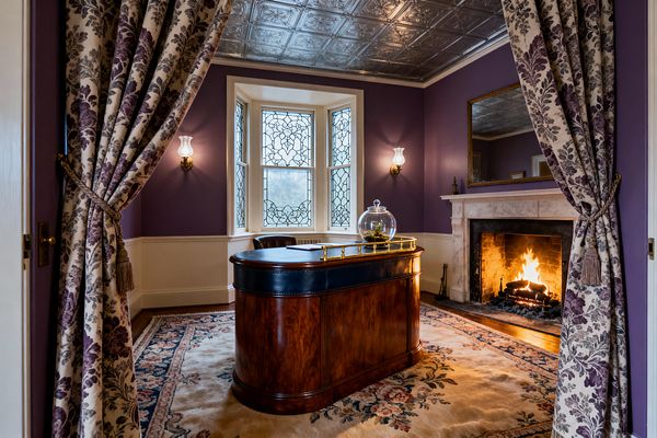 Victorian-style home office viewed through tied-back floral drapes. A polished mahogany kidney desk with a black leather top sits on a large floral wool rug, facing a roaring fire within a carved marble mantel and gilded mirror. Walls are painted deep plum above cream wainscoting, with brass sconces and leaded-glass bay windows. A patterned silver pressed-tin ceiling and hardwood floor complete the room’s ornate, warmly lit atmosphere.