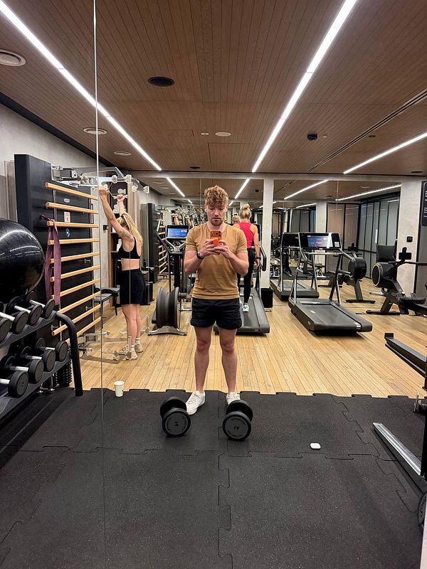 A man taking a mirror selfie in a modern gym with various workout equipment and other gym users.