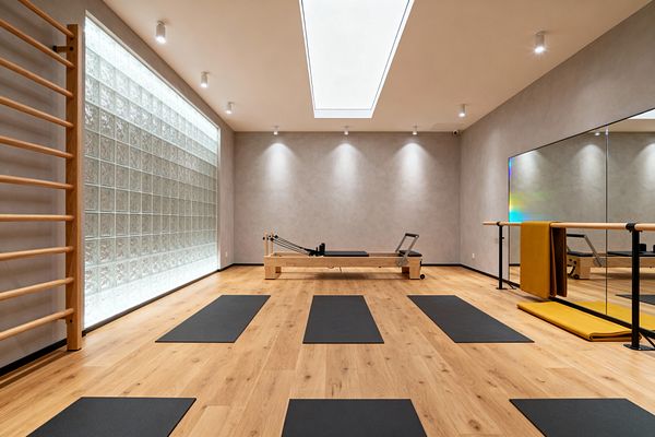 A minimalist home gym with light oak flooring features evenly spaced black mats facing a central wood Pilates reformer. The space has soft gray walls, a backlit glass-block wall with adjacent wooden stall bars, a full-width mirror and wood-and-black metal barre on the right, and a large rectangular skylight with recessed spotlights providing diffuse, even lighting, creating a calm, bright environment for stretching, barre, and Pilates work.
