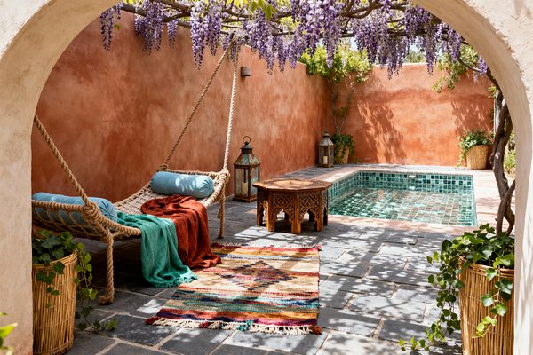 An intimate outdoor patio enclosed by sunbaked terracotta plaster walls features a small square plunge pool clad in glossy emerald zellige tiles. A thick-rope hanging daybed with pale blue bolster pillows holds teal and rust throws beside an intricately carved wooden coffee table. Lavender wisteria cascades from a pergola, casting dappled shade onto charcoal slate pavers, a colorful kilim rug, rattan planters with trailing greenery, and metal lanterns.