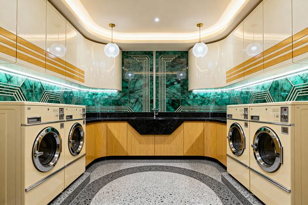 A symmetrical Art Deco laundry room features pairs of cream front‑loading machines with chrome trim flanking oak base cabinets and a black stone sink. An emerald marble backsplash with brass geometric inlays runs under glossy cream, curved upper cabinets with wood banding and LED strips; globe pendants and cove lighting illuminate a gray terrazzo floor with dark arcs, creating a glamorous, streamlined look.