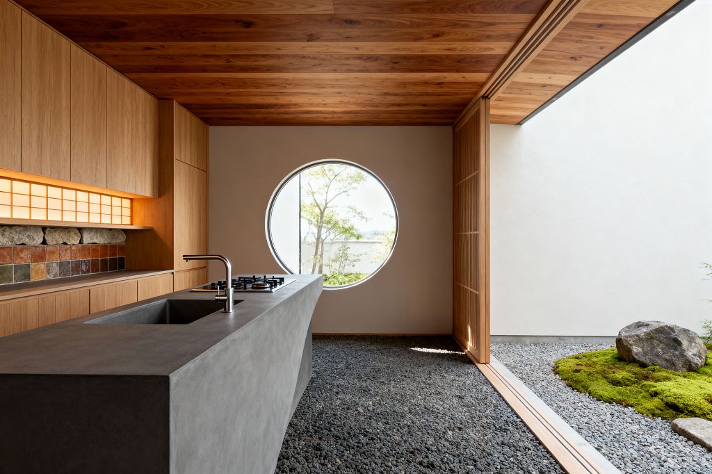 A minimalist Japanese Zen kitchen features a monolithic charcoal concrete island with integrated sink and black gas burners, set over a floor of dark river pebbles. Natural wood cabinetry and ceiling, a backlit shoji panel, and an earthy stone-and-ceramic tile backsplash frame a round window, while sliding wood lattice doors open to a gravel courtyard with a moss mound and boulder, bringing soft daylight and garden views into the serene space.