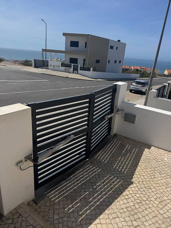 A damaged electric gate is shown in front of a modern house with a view of the ocean.