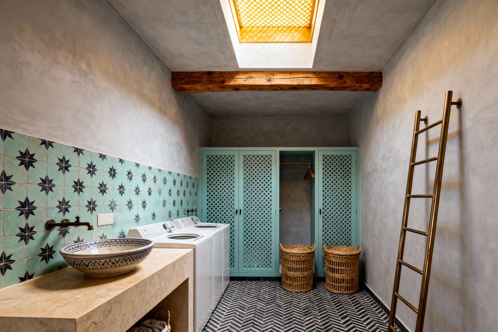 A Moroccan-style laundry room with soft gray plaster walls features a mint zellige tile backsplash patterned with navy starbursts. A beige travertine console holds a blue-patterned ceramic vessel sink and a wall-mounted brass faucet. A white washer and dryer sit below the tiles. Opposite, mint lattice-front cabinets and an open hanging nook stand beside two woven rattan hampers. A brass wall ladder, black-and-white chevron cement floor, timber beam, and a skylight with a perforated screen complete the space.