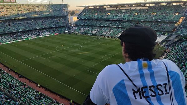 A fan wearing a Messi jersey watches a football match at a stadium.