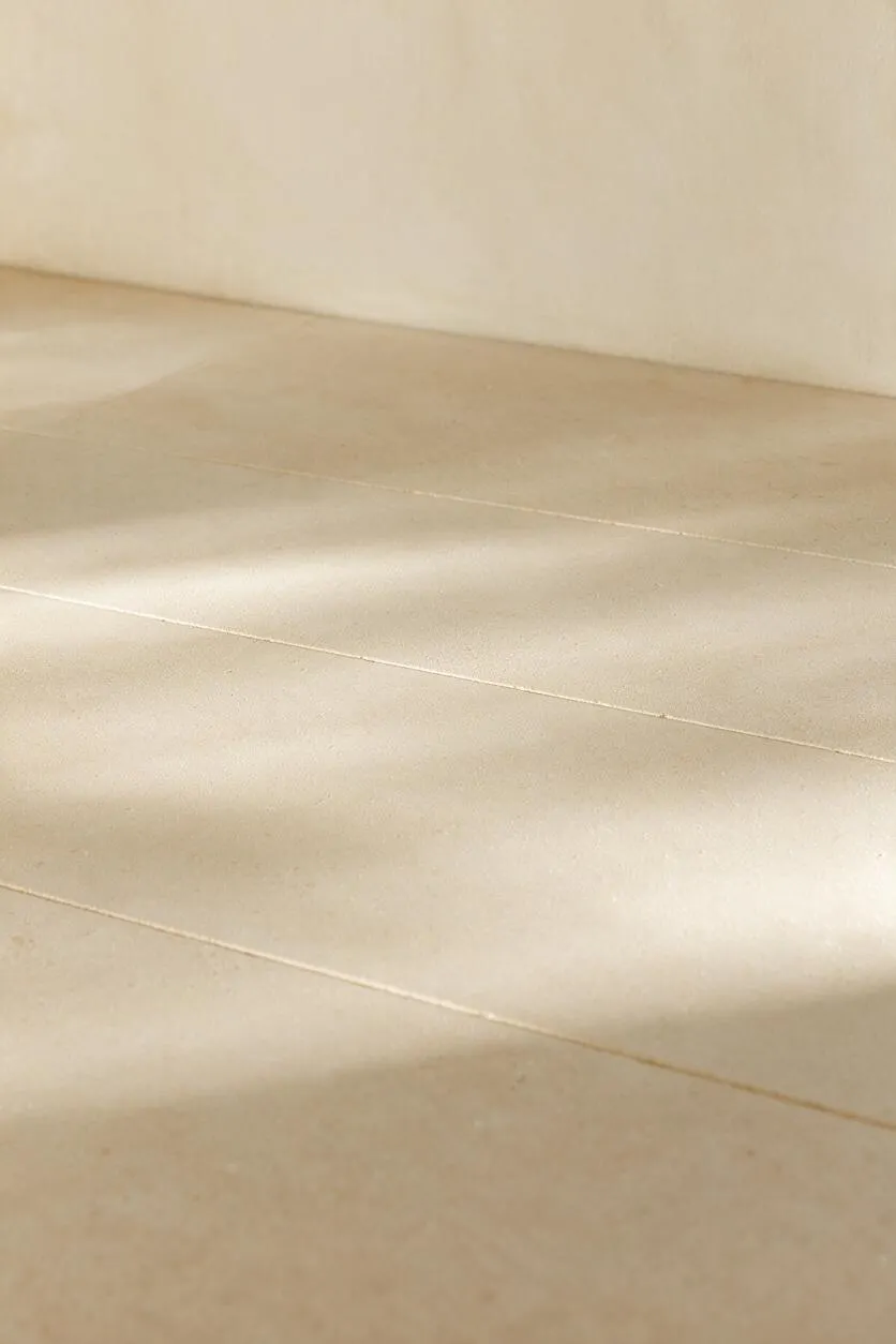A close-up view of a minimalist interior floor made of large beige stone or concrete tiles with thin grout lines. Soft natural light creates gentle highlights and shadows across the smooth surface, and a plain light wall meets the floor at the top of the image.